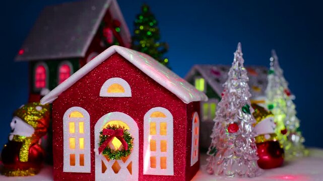 A close-up of a rotating Christmas town with houses with bright lights in the windows with green and glass Christmas trees and snowmen on a dark blue background