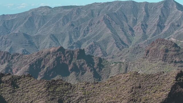 Aerial drone shot captures a massive, rugged mountain range with deep ridges and valleys, showcasing the dramatic volcanic landscape of Tenerife, Spain.