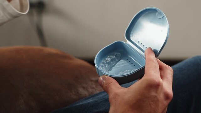 Close-up of hands opening a small blue case and removing a clear dental aligner. The scene highlights modern orthodontic treatment, oral care, personal hygiene, and at-home dental routines.