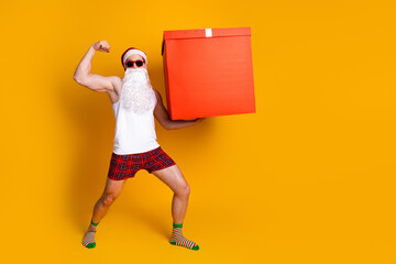 Santa in a white tank top red plaid shorts and striped socks carries a large red gift box against a bright yellow backdrop for a festive humorous holiday scene