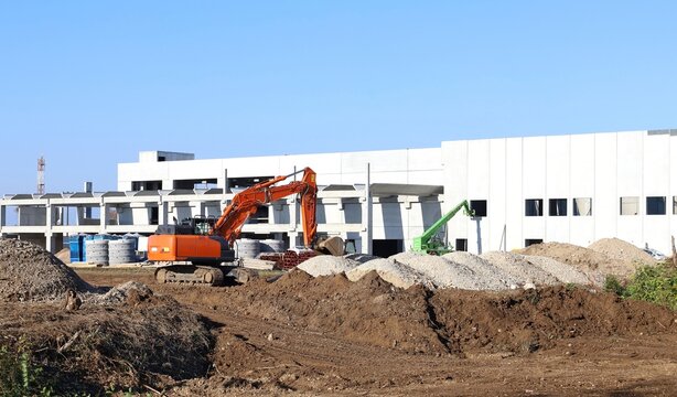 Heavy excavator and telescopic handler in front of a large industrial building under construction. Background for copy space.