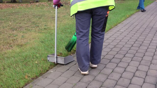 A sanitation worker in uniform and reflective vest walks along a paved sidewalk carrying a green broom and metal dustpan near a grassy edge