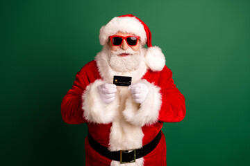 Santa in red suit with white fur holds a card wearing a hat and sunglass against a green background for christmas shopping and holiday promotion