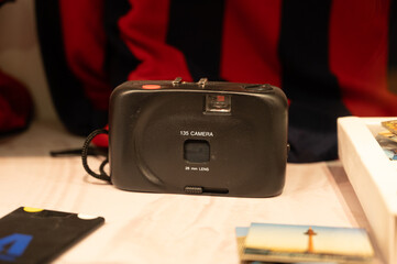Close-up of a black, simple 135 film camera (28mm lens) on a desk surrounded by old photos and photography accessories, evoking 90s nostalgia.