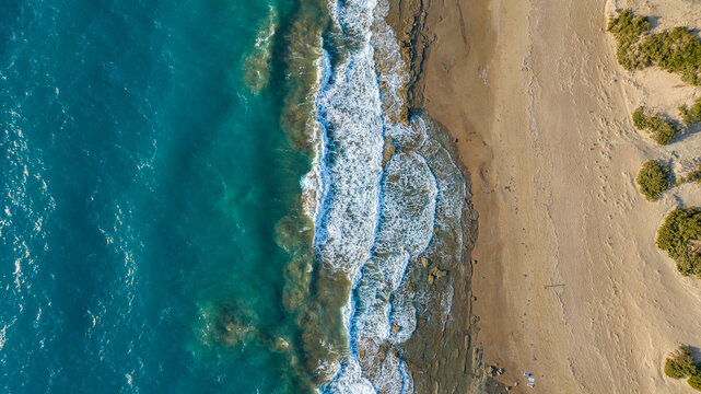Aerial view of turquoise waves crash against the sandy shore of the Aegean Sea, where golden sands meet the deep blue waters, Rhodes, Greece.