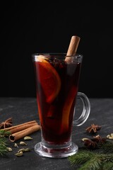 Tasty mulled wine in glass mug, spices and fir branches on black table, closeup