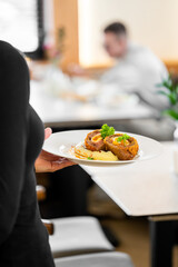 A close-up shot of a waiter holding a white plate with two gourmet, golden-brown meat rolls stuffed with egg, served on mashed potatoes in a restaurant setting