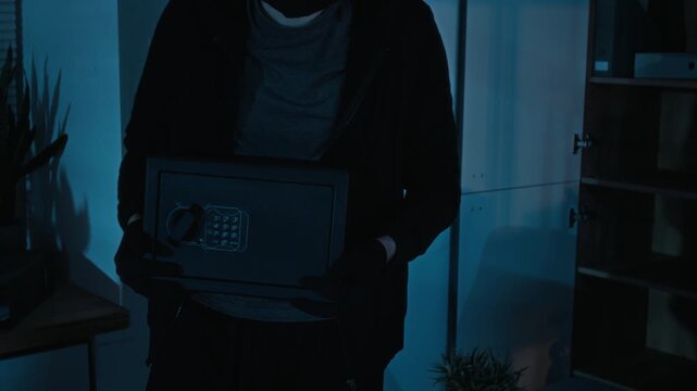 Midsection shot of male robber wearing black balaclava, hoodie and gloves stealing electronic safe from office cabinet during night break-in