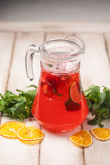 Red citrus lemonade in a glass pitcher with mint, orange slices and fresh herbs on rustic wooden background