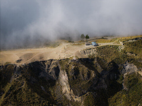 Aerial view of the crater lake's rim, where a small structure and a pair of trees stand against the backdrop of low-hanging clouds, Quilotoa lake, Cotopaxi province, Pujili Canton, Ecuador.