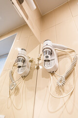 Wall Mounted Hair Dryer in Beige Tiled Bathroom with Coiled Cords and Mirror. Hotel or spa focused on guest grooming and convenience.