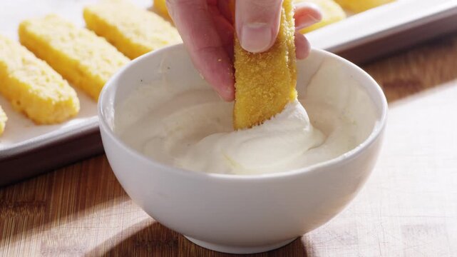 A hand dips a crispy fish stick into a bowl of thick white sauce . High quality 4k footage