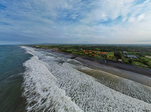 Aerial view of the dark sands meeting the frothy waves in Seminyak, where resorts nestle amidst the lush greenery, Seminyak, Bali, Indonesia.