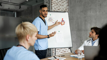Middle-eastern young man doctor showing his colleagues presentation, multiracial team of professional docotrs attending medical conference or having breefing, discussing medical cases, copy space