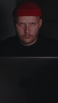 Vertical close-up from behind the laptop lid: a man lifts his eyes toward the camera with a strict, focused expression. An intense and concentrated moment in a dark tech workspace. High quality 4k