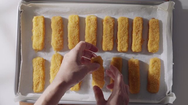 A pair of hands prepares breaded fish sticks on a parchment-lined baking tray, flat lay. High quality 4k footage