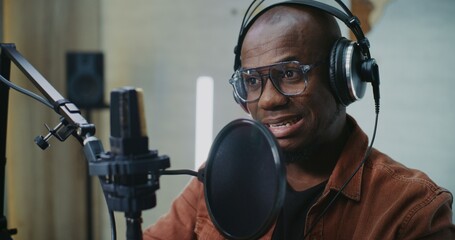 Confident African American Podcast Guest Wearing Glasses and Large Headphones Speaks Thoughtfully Into Pro Microphone With Pop Filter, Smiling as Engages in Meaningful Studio Conversation. Portrait.