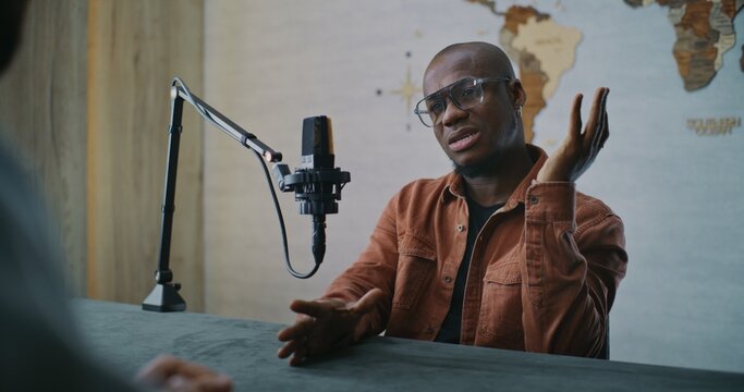 Confident Podcast Guest Engaged in Thoughtful Studio Discussion. African American Man Participates in Mindful Dialogue, Smiling as Responds to Host Across Table in Modern Podcast Studio. Portrait. - Powered by Adobe