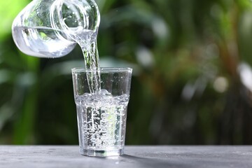 Pouring water into glass from jug at grey table against blurred green background, closeup. Space for text