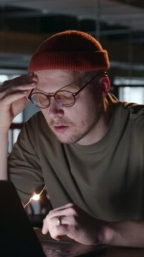 Vertical close-up video of a man leaning toward his laptop with his hand on his head, struggling with a difficult task or complex problem. A tense moment of concentration in a dim workspace. 4k