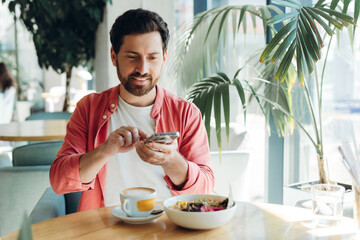 Happy handsome Latin bearded man holding mobile phone, taking photo of food