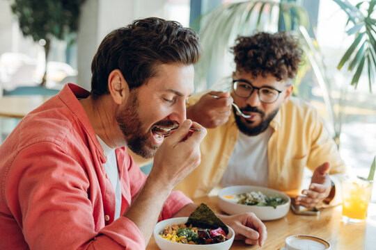 Smiling, happy diverse men wearing casual clothes eating in modern cafe, holding spoon