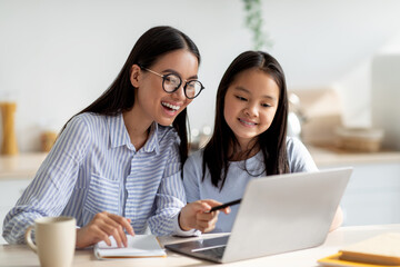 A joyful Asian mother and her daughter are sitting in the kitchen, focused on a laptop screen. The girl is engaged in online learning while the mother offers guidance and support.