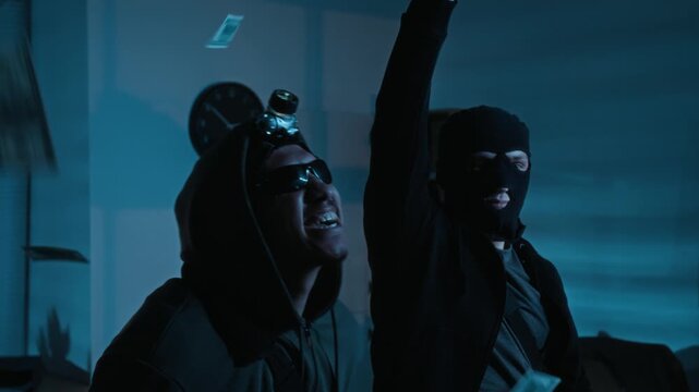 Two delighted young robbers throwing dollar bills in air celebrating successful burglary in dimly lit office