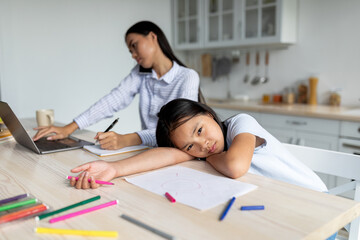 Young korean mother is busy working from home with her laptop, ignoring her little girl who feels offended and lonely. The daughter sits at the table feeling abandoned, waiting for attention.