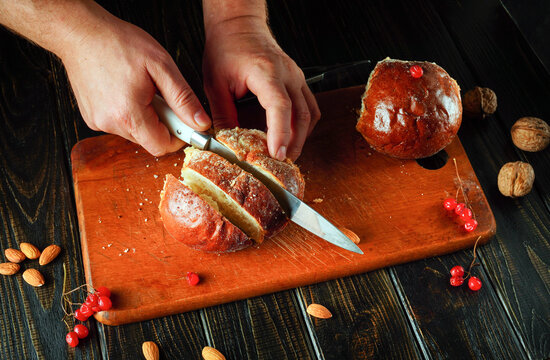 Hands carefully slice a round loaf of bread or bun on a wooden cutting board. Almonds, walnuts, and red berries are arranged around the loaf, creating a rustic kitchen scene