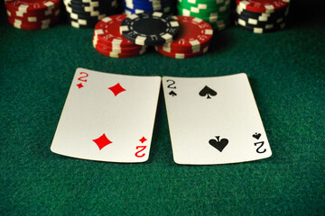 Two cards showing the 2 of diamonds and 2 of spades are placed on a green felt poker table. In the background, various colorful poker chips are stacked high, creating an exciting casino atmosphere