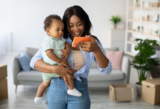Smiling African American mother holds her little son while enjoying a video call at home. They are in a cozy living room filled with boxes, sharing laughter over the phone.