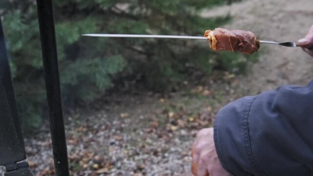 Close-up of male hands threading chunks of marinated raw meat onto a sharp metal skewer. The cook prepares shish kebabs outdoors with spicy orange marinade coating his fingers.