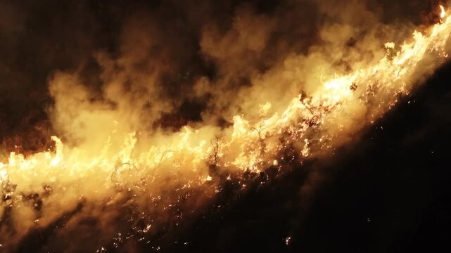Aerial view of a wildfire blazing across the landscape, smoke billowing into the air, creating a stark contrast between the orange flames and dark background, South Africa.