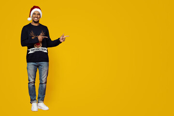 Naklejka premium A man wearing a Christmas sweater and a Santa hat stands smiling and pointing to the left. The background is a vibrant yellow, adding to the cheerful holiday mood.