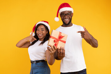 A joyful African American couple is pointing at a wrapped Christmas present while smiling. They wear Santa hats and are set against a bright yellow studio background, celebrating the holiday spirit.