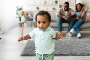 Toddler joyfully takes first steps in a cozy living room. Parents encourage him from the sofa,...