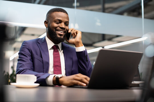 Smiling black man in suit manager working on laptop and talking on mobile phone at office, drinking coffee, successful businessman having phone conversation with clients, checking data online