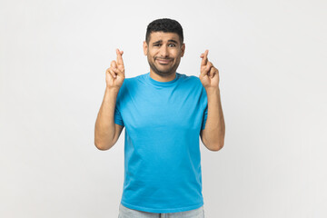 Unshaven man standing keeps fingers crossed, hope for good luck, frowning face, looking at camera.