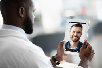 Side view of bearded black man in white robe doctor having video call with male patient, using...