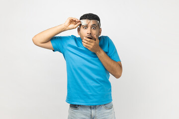 Shocked man raised his optical eyeglasses, sees something astonished, covering mouth with palm.