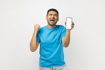 Joyful positive man showing blank white screen of cell phone, clenches fist, rejoicing his success.