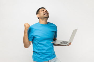 Extremely happy man holding laptop in hands, clenches fist, betting and winning celebrating success