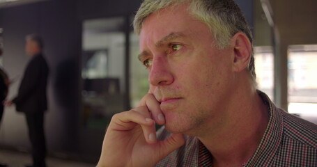 Close up of middle aged man with thoughtful expression, hand resting on chin, gazing forward in silence, seated in office environment with blurred colleagues in background