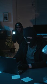 Vertical view of cyber thief in black balaclava stealing personal data from laptop in office, accomplice going up to him with torch and asking about progress