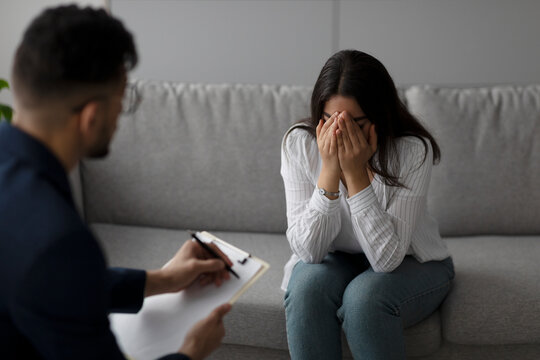 An Arab woman sits on a sofa in a clinic, crying as she consults with a psychologist. She is letting out her negative emotions and experiencing a nervous breakdown during the session.