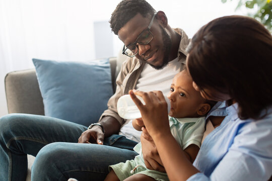 Loving African American parents spend time nurturing their cute toddler. They smile as they feed their baby boy from a bottle, enjoying a joyful moment together on the couch. - Powered by Adobe