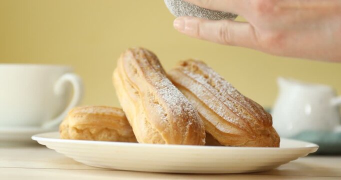 Woman sprinkling eclairs with powdered sugar at white wooden table, closeup