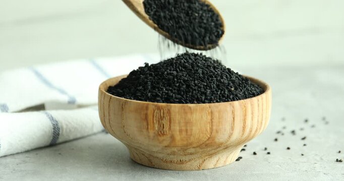 Woman pouring aromatic black caraway into wooden bowl at grey table, closeup