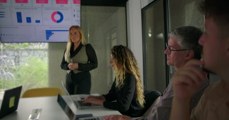 Businesswoman standing in front of screen while explaining data points to engaged team seated around table in modern office setting